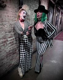 Two costumed performers in black-and-white striped outfits pose against a textured brick hallway: one in white face paint, feathered headpiece and harlequin skirt, the other with green hair, black hat and striped pants playfully pulling a tie.