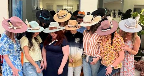 Group of women indoors trying on colorful wide-brim hats at a playful hat-try-on gathering, casual outfits and floral dresses visible