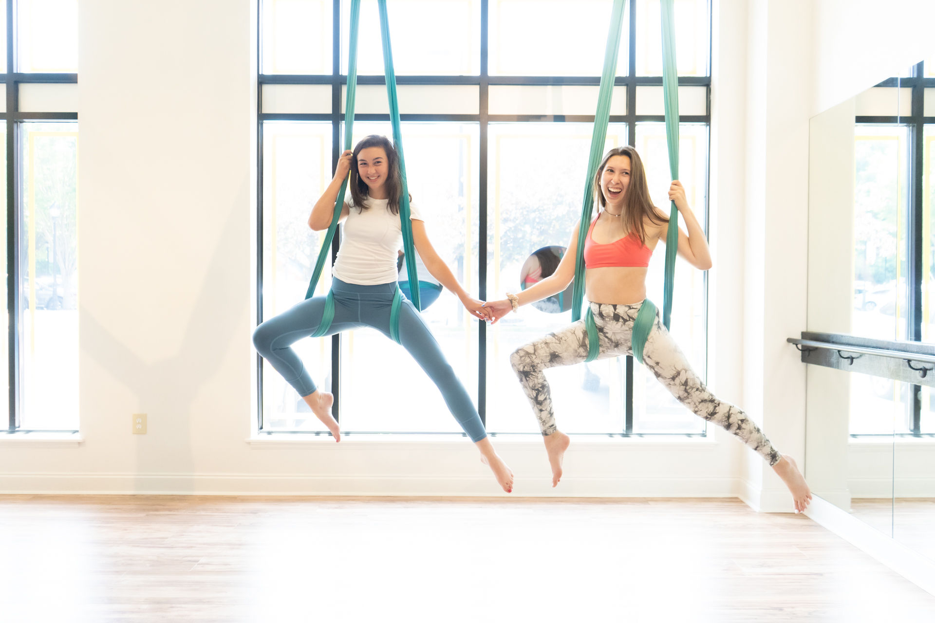 Two women smiling in a sunlit aerial yoga studio, suspended in green hammocks holding hands mid-pose by large windows and hardwood floors.
