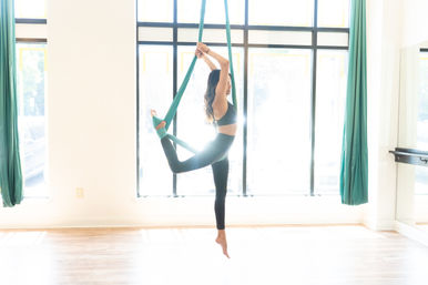Woman practicing aerial yoga on teal silks in a bright sunlit studio, balancing with one leg wrapped in fabric and arms reaching overhead.