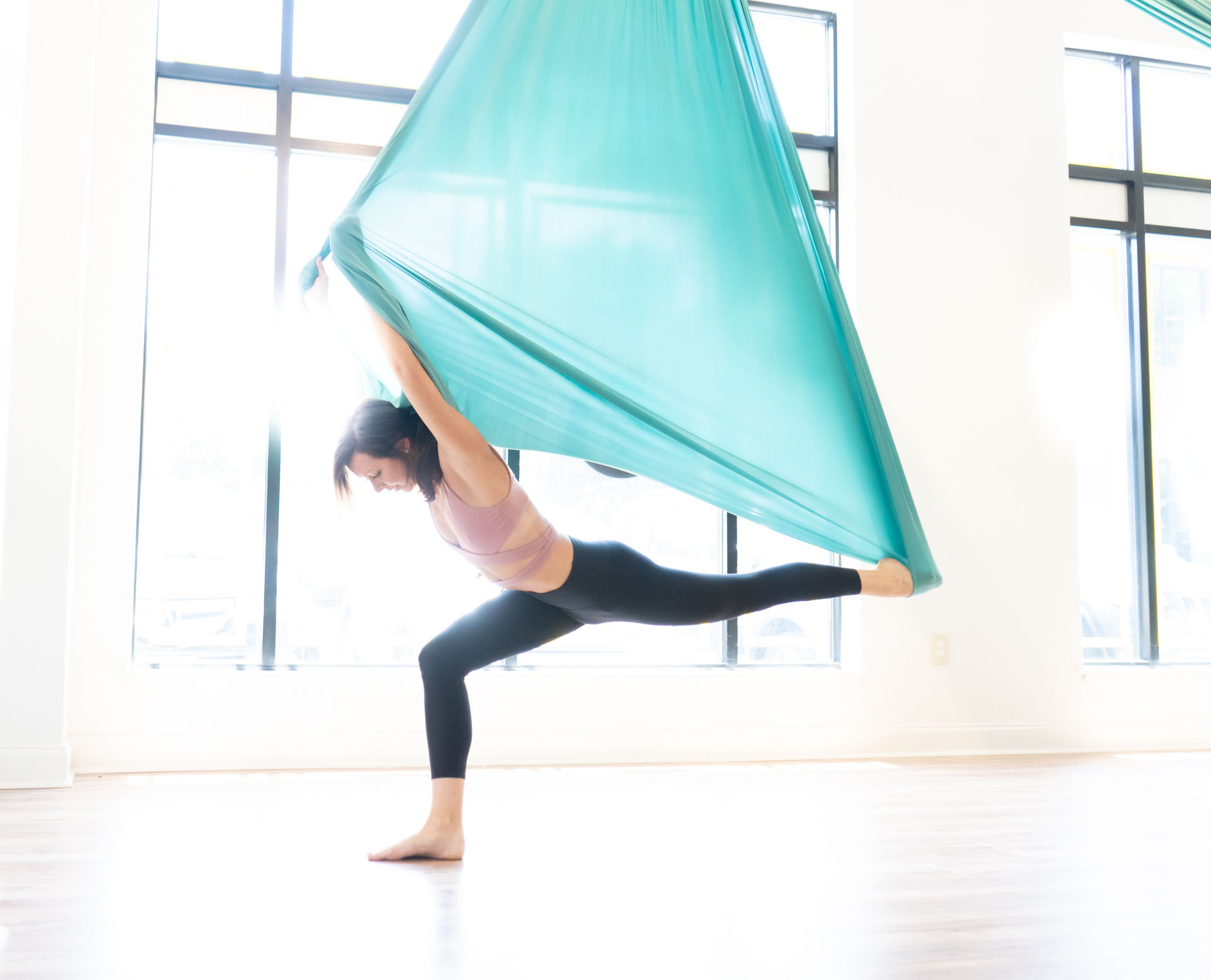 Woman performing aerial yoga split in a teal silk hammock, balancing on one leg in a bright sunlit studio with large windows.