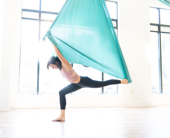 Woman performing aerial yoga split in a teal silk hammock, balancing on one leg in a bright sunlit studio with large windows.