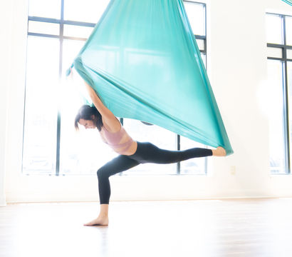 Woman performing aerial yoga split in a teal silk hammock, balancing on one leg in a bright sunlit studio with large windows.