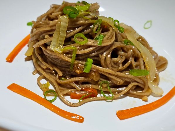 Neatly twirled Japanese buckwheat soba noodles on a white plate, tossed with sautéed onions, thin carrot strips and sliced scallions.