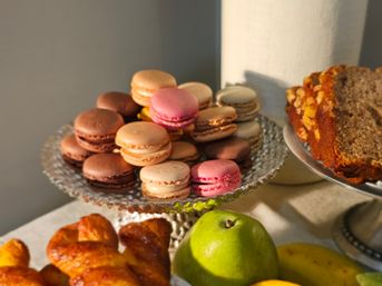 Colorful assorted macarons piled on a glass pedestal cake stand beside croissants, a green pear, bananas and a nut-topped banana bread slice on a dessert table