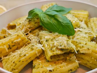 Close-up of rigatoni pasta coated in creamy green pesto, sprinkled with grated Parmesan and garnished with fresh basil leaves in a white bowl.