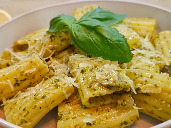 Close-up of rigatoni pasta coated in creamy green pesto, sprinkled with grated Parmesan and garnished with fresh basil leaves in a white bowl.