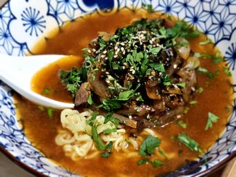 Close-up of beef ramen: curly noodles in rich brown broth topped with braised onions, sesame seeds and chopped fresh herbs, served in a blue-and-white patterned bowl with a porcelain spoon.