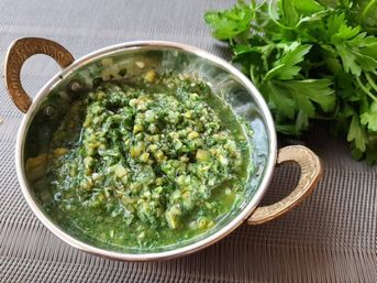 Fresh chimichurri-style parsley and garlic sauce in a small metal serving bowl with ornate brass handles on a placemat, chopped herbs and garlic visible and a bunch of fresh parsley beside it.