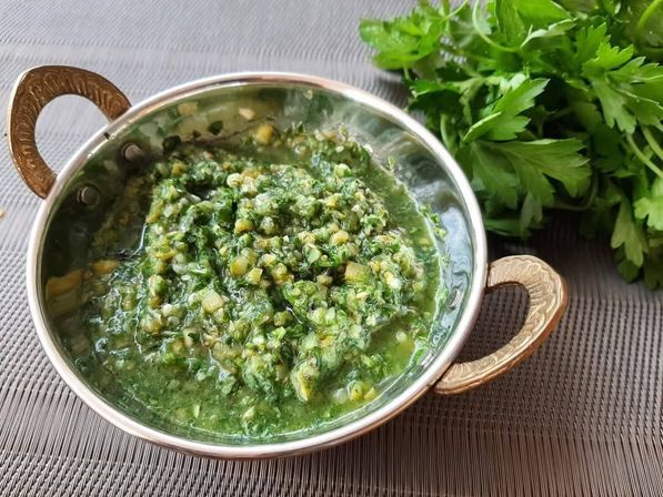 Fresh chimichurri-style parsley and garlic sauce in a small metal serving bowl with ornate brass handles on a placemat, chopped herbs and garlic visible and a bunch of fresh parsley beside it.