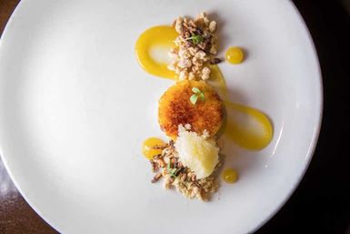 Fine‑dining plated dessert: caramelized custard with bright mango‑citrus purée, crunchy crumble and a scoop of sorbet on a white plate.