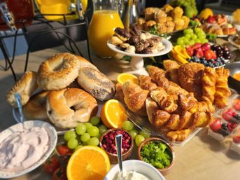 Vibrant brunch buffet with poppy-seed bagels, golden croissants and pastries, muffins and cookies, bowls of green grapes, strawberries, blueberries and pomegranate seeds, halved oranges, cream cheese, yogurt and a pitcher of orange juice.