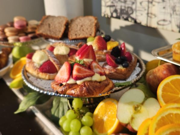 Colorful brunch spread: strawberry-topped cream pastries and danishes on a tiered tray with macarons, sliced oranges and apples, and green grapes.