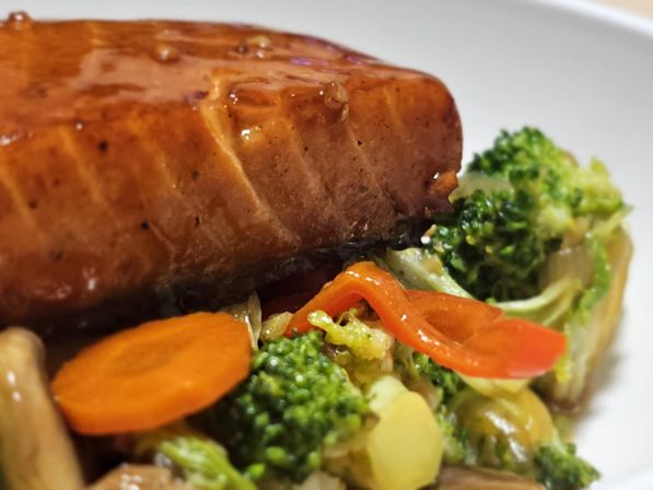 Close-up of seared glazed salmon fillet atop sautéed broccoli, sliced carrots and red pepper on a white dinner plate