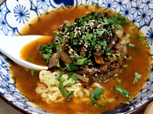 Hearty Japanese-style beef ramen with curly noodles in rich miso broth, topped with braised beef, caramelized onions, sesame and chopped cilantro in a blue patterned bowl