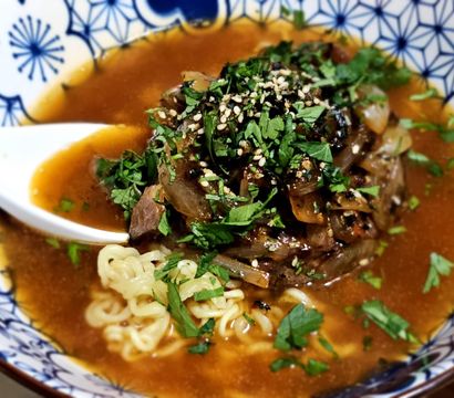 Hearty beef ramen in a blue patterned bowl — curly noodles in a rich amber broth topped with braised beef, caramelized onions, sesame seeds and chopped fresh herbs with a white soup spoon
