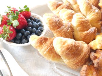 Flaky golden croissants on a platter beside a bowl of fresh strawberries and blueberries on a light linen table — bright breakfast/brunch close-up.