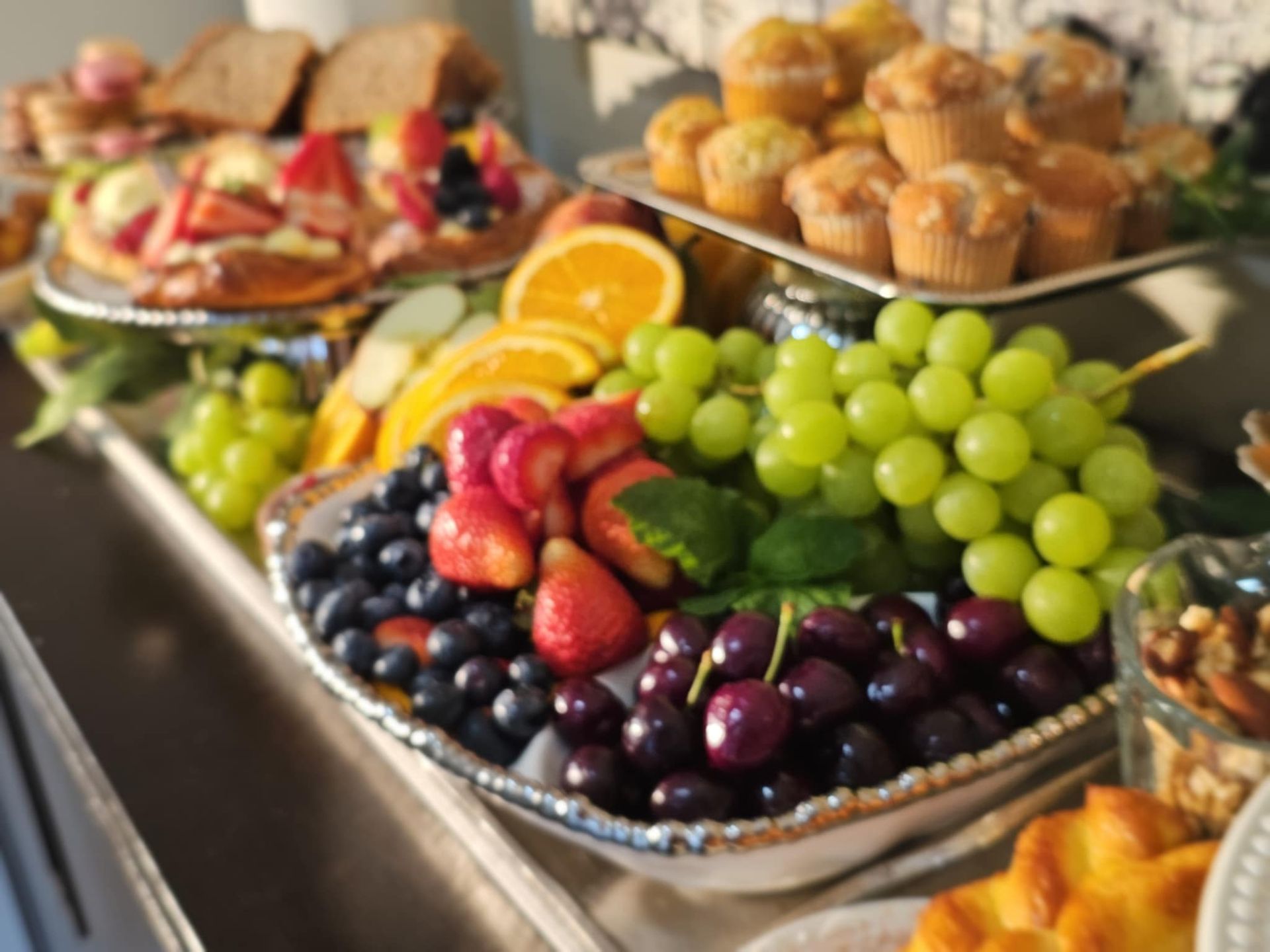 Colorful brunch buffet with fresh green grapes, cherries, strawberries, blueberries and orange slices on a silver platter, surrounded by muffins and assorted pastries