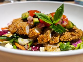 Crispy breaded chicken slices atop a colorful mixed greens salad with cherry tomatoes, cucumber, red onion, feta and fresh basil in a white bowl