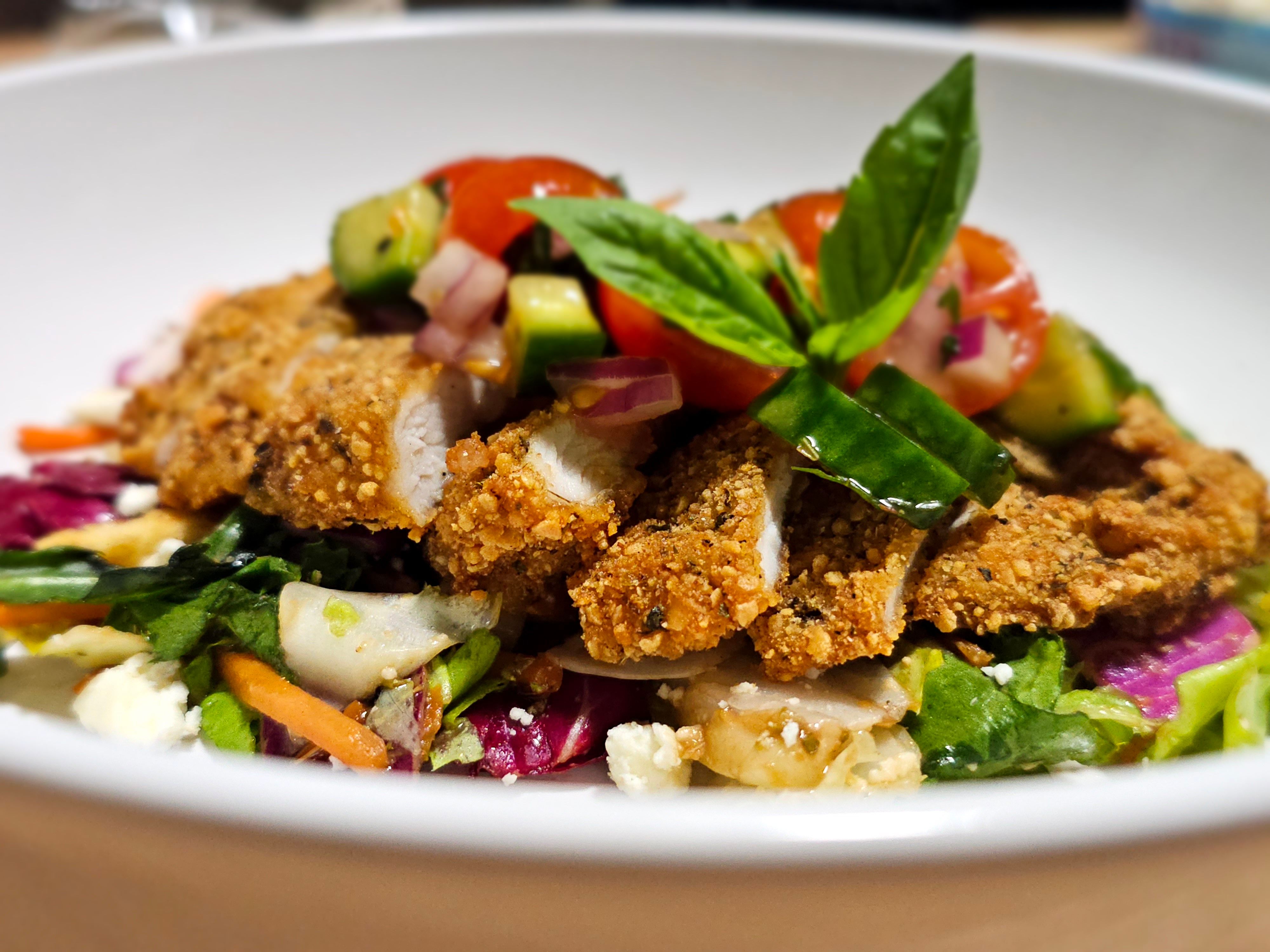 Close-up of crispy breaded chicken slices on mixed greens with cherry tomato and cucumber salsa, fresh basil and feta in a white bowl