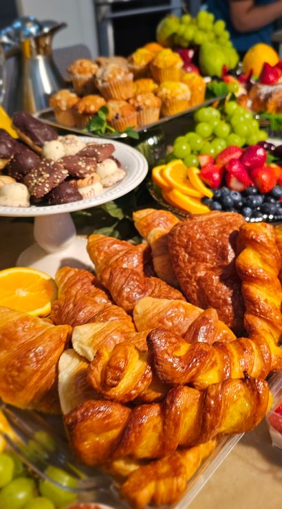 Brunch buffet table with flaky croissants and twisted pastries, poppy seed bagels, chocolate-dipped cookies, mini yogurt and berry parfaits, mixed fruit platter with grapes, strawberries and orange slices, macarons, muffins and a pitcher of orange juice