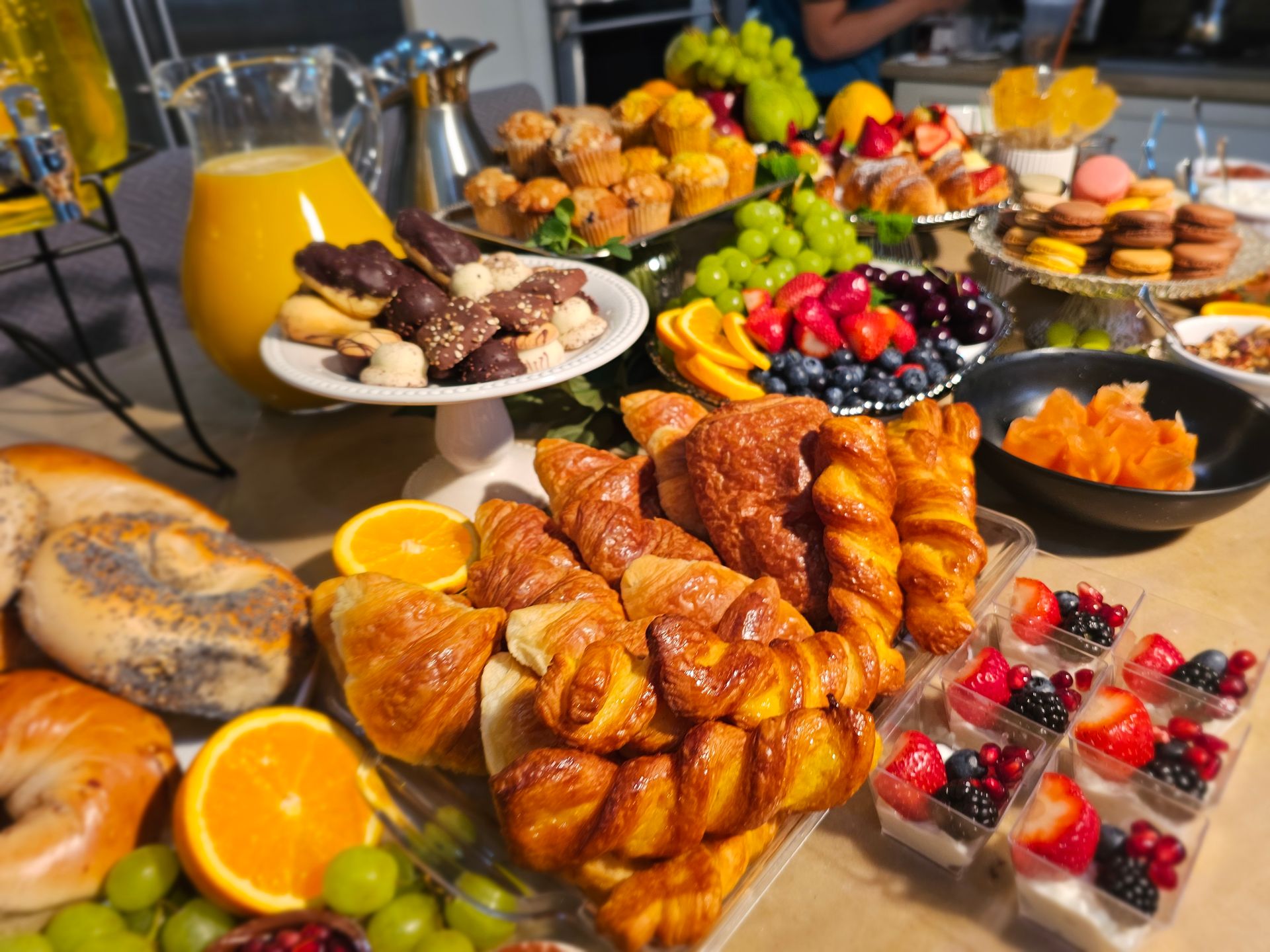 Brunch buffet table with flaky croissants and twisted pastries, poppy seed bagels, chocolate-dipped cookies, mini yogurt and berry parfaits, mixed fruit platter with grapes, strawberries and orange slices, macarons, muffins and a pitcher of orange juice