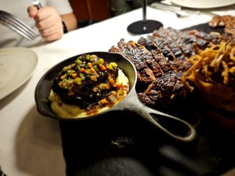 Hearty steakhouse platter: cast-iron skillet of creamy mashed potatoes topped with braised short rib and diced vegetables, sliced grilled steak and crispy onion strings on a white-clothed table.