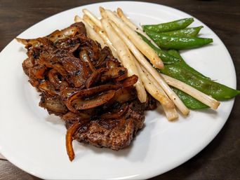 Close-up of grilled steak topped with caramelized onions, served with white asparagus spears and bright sugar snap peas on a white plate over a wooden table.