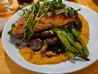 Grilled salmon fillet topped with microgreens and crispy onions, served on roasted baby potatoes and charred asparagus over a bright carrot–sweet potato puree with brown sauce on a white plate.