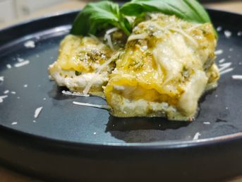 Close-up of Italian-style cheesy baked pasta squares with pesto, oozy melted cheese, grated Parmesan and fresh basil on a black plate.