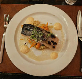Seared salmon fillet with crispy skin atop quinoa and roasted carrots, topped with microgreens and surrounded by four dollops of creamy aioli on a white dinner plate in a restaurant setting.