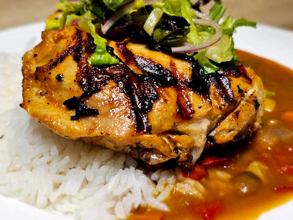 Juicy grilled chicken thigh with charred grill marks on steamed white rice, served with savory brown vegetable sauce and topped with shredded lettuce and red onion — close-up plate shot