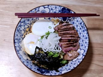 Overhead shot of a Japanese-style ramen bowl on a wooden table with soft-boiled eggs, sliced seared duck, shredded nori, scallions and pickled white onion in a blue patterned bowl with chopsticks.