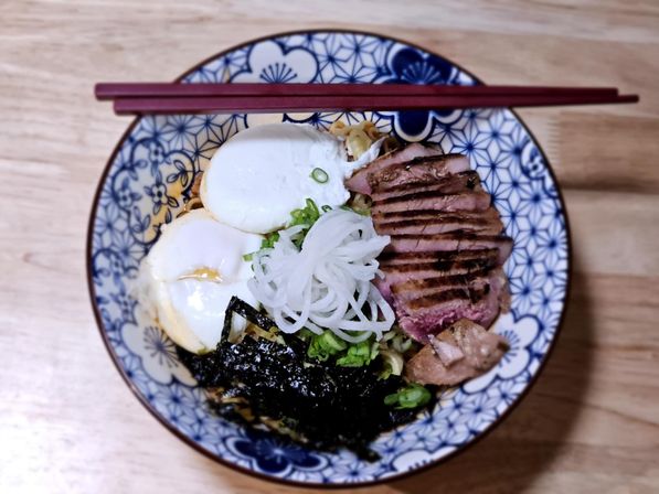 Overhead shot of a Japanese-style ramen bowl on a wooden table with soft-boiled eggs, sliced seared duck, shredded nori, scallions and pickled white onion in a blue patterned bowl with chopsticks.