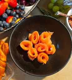 Black bowl with six smoked-salmon roses on a brunch spread surrounded by fresh blueberries, strawberries, green grapes and a croissant