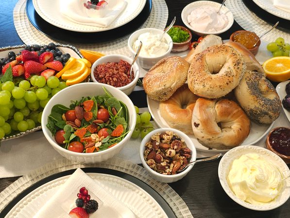 Inviting brunch spread on a dining table with a stack of assorted bagels (sesame, poppy, plain), bowls of cream cheese and butter, mixed green salad with cherry tomatoes and nuts, fresh fruit platter with grapes, strawberries, blueberries and orange slices, plus granola, jam and toppings.