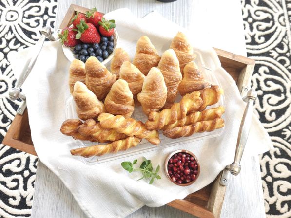 Overhead view of a wooden breakfast tray with golden mini croissants and twisted pastry sticks, a bowl of strawberries and blueberries, and a small dish of pomegranate seeds on white linen
