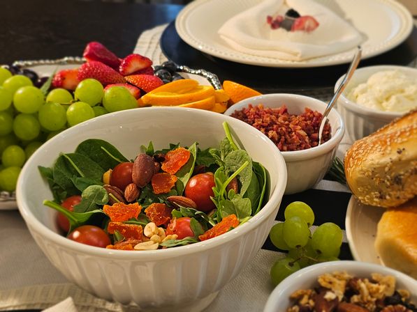 Brunch table with a white bowl of spinach salad topped with cherry tomatoes, nuts and dried apricots, surrounded by green grapes, sliced strawberries and oranges, small bowls of bacon bits and cream cheese, and sesame bagels.
