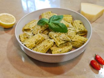 Bowl of pesto-coated rigatoni pasta topped with grated Parmesan and a fresh basil sprig, lemon half, cheese wedge and cherry tomatoes on a beige countertop