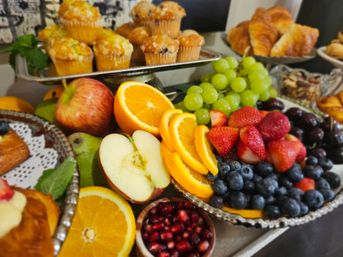 Colorful brunch buffet display on silver trays with sliced oranges and apple halves, green grapes, strawberries, blueberries and pomegranate seeds, surrounded by muffins and croissants