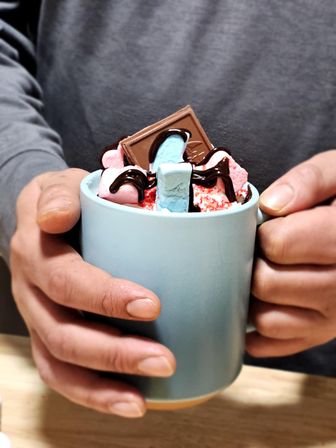 Hands holding a blue ceramic mug of gourmet hot chocolate topped with colorful marshmallows, a chocolate square, chocolate drizzle and red sprinkles