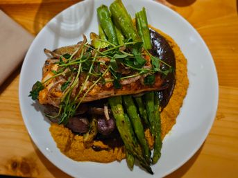 Grilled salmon fillet on roasted asparagus and potatoes over a bright carrot purée with dark glaze, topped with microgreens on a white plate on a wooden table