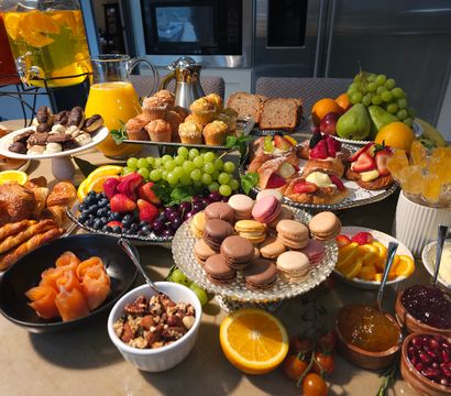 Vibrant brunch spread on a kitchen island with croissants, muffins, assorted macarons, fruit platters (grapes, strawberries, blueberries, orange slices), smoked-salmon roses, berry-topped pastries, sliced bread, mixed nuts, jams, yogurt, and pitchers of orange juice and beverages.