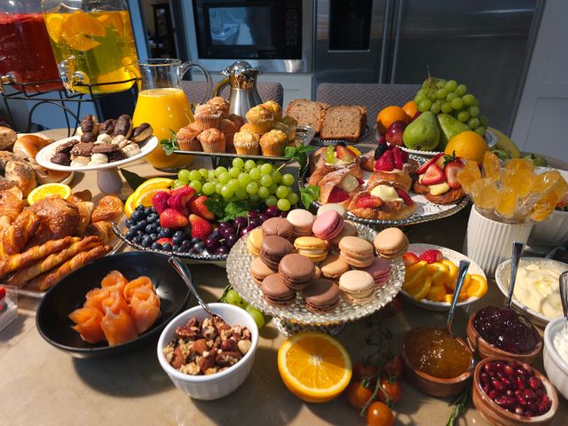 Vibrant brunch spread on a kitchen island with croissants, muffins, assorted macarons, fruit platters (grapes, strawberries, blueberries, orange slices), smoked-salmon roses, berry-topped pastries, sliced bread, mixed nuts, jams, yogurt, and pitchers of orange juice and beverages.