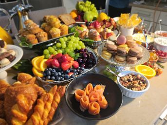 Colorful brunch spread on a kitchen island with croissants and twisted pastries, muffins, macarons, fresh fruit platter (grapes, strawberries, blueberries, orange slices, cherries), smoked salmon rosettes, mixed nuts and jams.