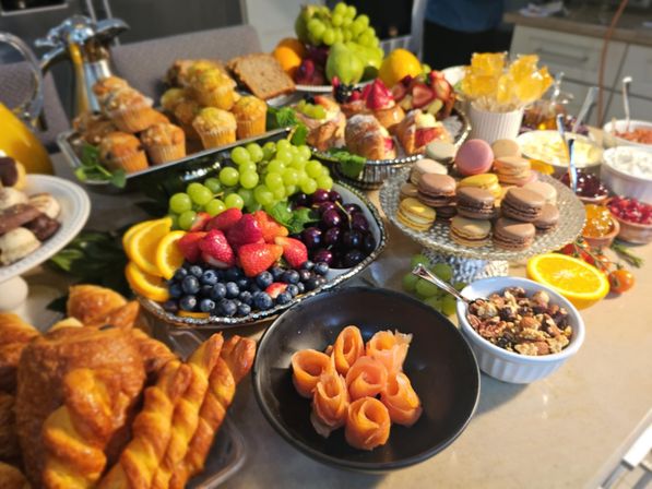 Gourmet brunch spread on a kitchen island: croissants and twisted pastries, muffins, a fruit platter with grapes, strawberries, blueberries and orange slices, a tier of colorful macarons, smoked-salmon rosettes, mixed nuts and jams.