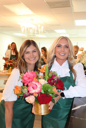 Two smiling women in green aprons holding a colorful bouquet in a gold vase at a bright floral-arranging workshop studio, chandeliers overhead and people arranging flowers in the background.