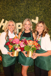 Three smiling women in green aprons hold vibrant mixed-flower bouquets in gold vases against a boxwood green wall with gold balloon letters, at a flower-arranging workshop.