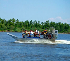 Airboat tour speeding across a river with passengers on board, cutting a white wake past tree-lined wetlands under a bright blue sky.