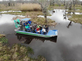 Aerial view of a blue-green airboat carrying a group through cypress swamp wetlands with marsh grasses and reflective water, Everglades-style